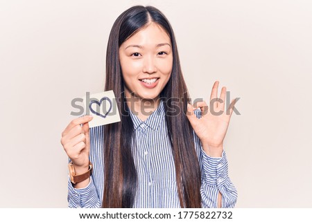 Young beautiful chinese woman holding reminder with heart shape doing ok sign with fingers, smiling friendly gesturing excellent symbol 