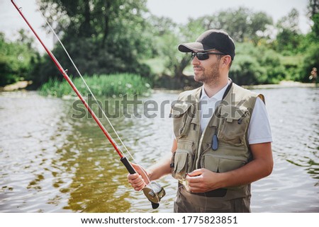 Young fisherman fishing on lake or river. Picture of serious professional guy holding rod in hands and waiting for fish. Stands in river or lake water. Catching tasty delicious fish.