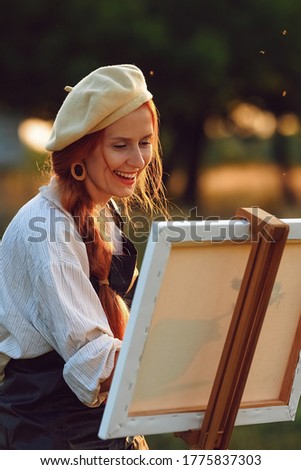 a girl artist with long red hair draws on an easel with a brush against the background of the sunset