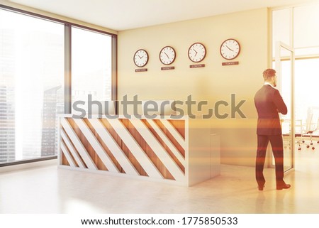 Confident young businessman standing in international company office lobby with white and wooden reception and clocks showing world time. Blurry cityscape. Toned image