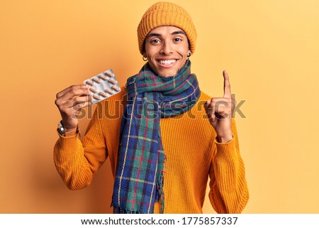 Young african amercian man wearing winter clothes holding pills smiling with an idea or question pointing finger with happy face, number one 