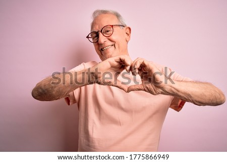 Grey haired senior man wearing glasses standing over pink isolated background smiling in love showing heart symbol and shape with hands. Romantic concept.