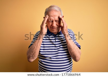 Grey haired senior man wearing casual navy striped t-shirt standing over yellow background with hand on head for pain in head because stress. Suffering migraine.