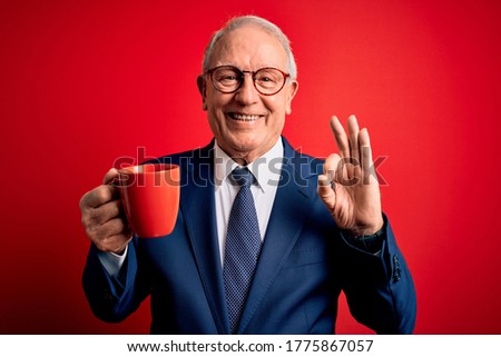 Senior grey haired business man drinking a hot cup of coffee over red background doing ok sign with fingers, excellent symbol
