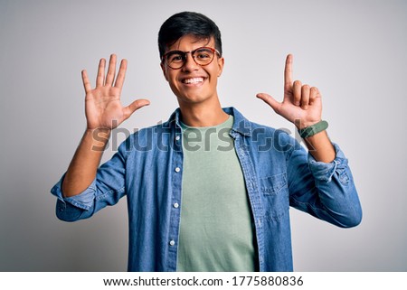 Young handsome man wearing casual shirt and glasses over isolated white background showing and pointing up with fingers number seven while smiling confident and happy.