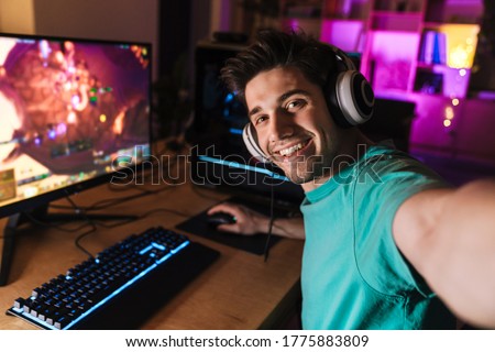 Image of caucasian cheerful man taking selfie photo while playing video game on computer at home