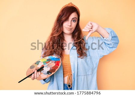 Young beautiful woman holding paintbrush and palette with angry face, negative sign showing dislike with thumbs down, rejection concept 