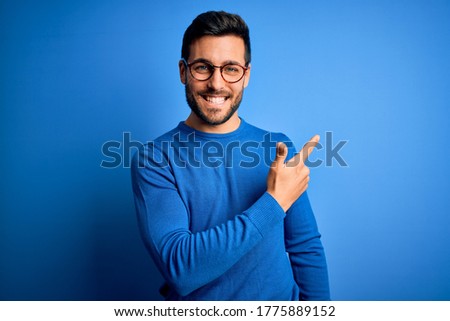 Young handsome man with beard wearing casual sweater and glasses over blue background cheerful with a smile on face pointing with hand and finger up to the side with happy and natural expression