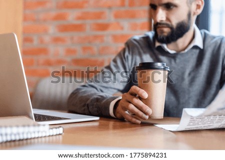 selective focus of handsome businessman looking at laptop while holding newspaper and paper cup