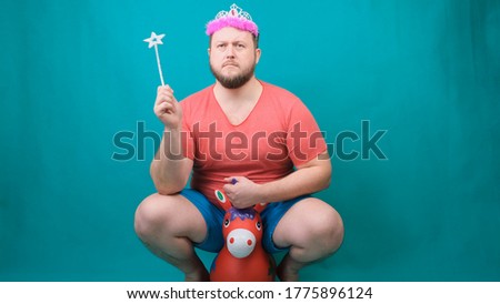 handsome bearded freaky man in a pink T-shirt with a deadema on his head is saddened riding a unicorn with a magic wand in his hand. A funny wizard joke to make and fulfill a wish