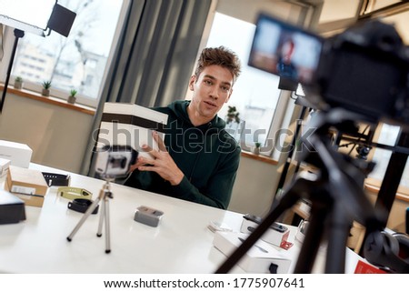 Young male technology blogger recording video blog or vlog about new camera lens and other gadgets at home studio. Blogging, Work from Home concept. Focus on person. Dutch angle. Horizontal shot Royalty-Free Stock Photo #1775907641