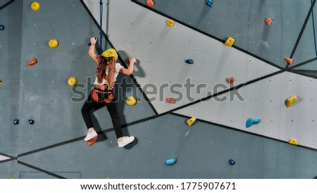 Girl in safety equipment and harness training on the artificial climbing wall indoors. Bouldering training concept. Rear view. Horizontal shot. Web Banner