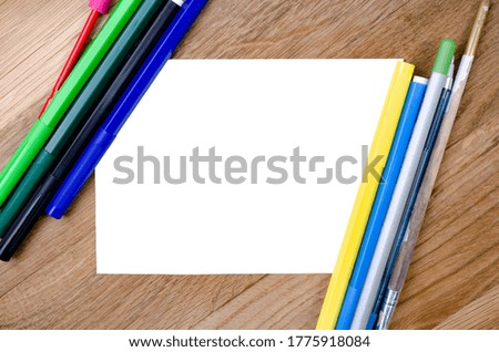 A white sheet of paper on a wooden background and stationery are nearby. Sheet of paper and pencils, felt-tip pens. Mock up