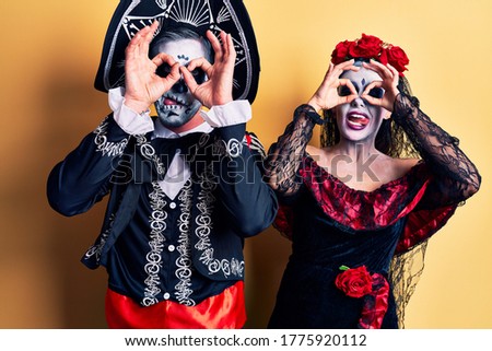 Young couple wearing mexican day of the dead costume over yellow doing ok gesture like binoculars sticking tongue out, eyes looking through fingers. crazy expression. 