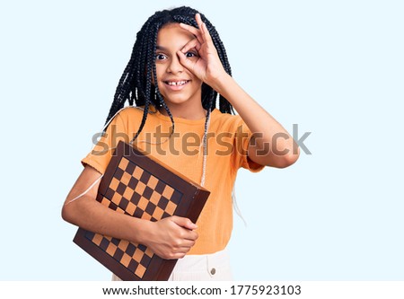 Cute african american girl holding chess smiling happy doing ok sign with hand on eye looking through fingers 