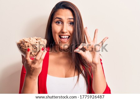 Young beautiful brunette woman holding bowl of cornflakes cereal doing ok sign with fingers, smiling friendly gesturing excellent symbol 