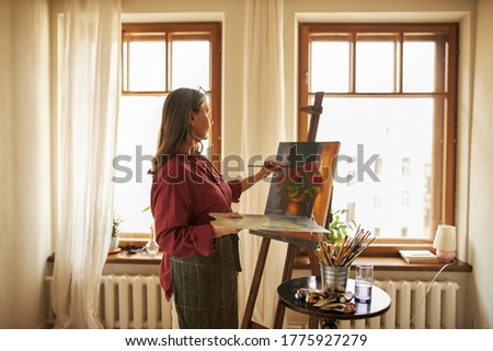 Side view of professional gray haired female artist on retirement standing in spacious room with large windows, holding palette, applying oil paint using brush, finishing beautiful picture on canvas