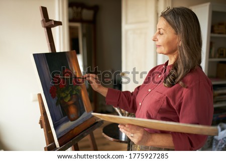Attractive stylish mature retired female having inspired look holding brush and palette painting flowers on canvas using oil paint, standing in front of easel during art class for beginners