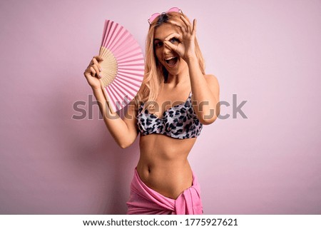 Young beautiful blonde woman on vacation wearing bikini using hand fan over pink background with happy face smiling doing ok sign with hand on eye looking through fingers