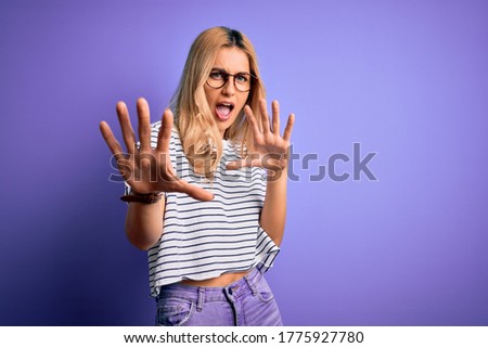 Young beautiful blonde woman wearing striped t-shirt and glasses over purple background afraid and terrified with fear expression stop gesture with hands, shouting in shock. Panic concept.