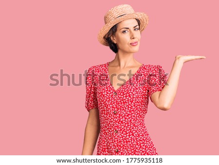Young beautiful woman wearing summer hat smiling cheerful presenting and pointing with palm of hand looking at the camera. 