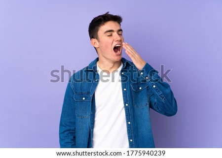 Teenager caucasian  handsome man isolated on purple background yawning and covering wide open mouth with hand