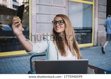 Cheerful elegant female in eyeglasses taking selfie with mobile phone and smiling while sitting in outdoor cafe in summer day