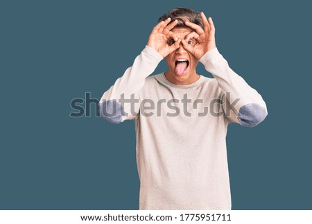 Young handsome man wearing casual clothes doing ok gesture like binoculars sticking tongue out, eyes looking through fingers. crazy expression. 