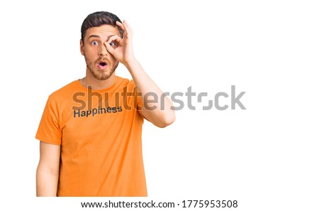 Handsome young man with bear wearing tshirt with happiness word message doing ok gesture shocked with surprised face, eye looking through fingers. unbelieving expression. 