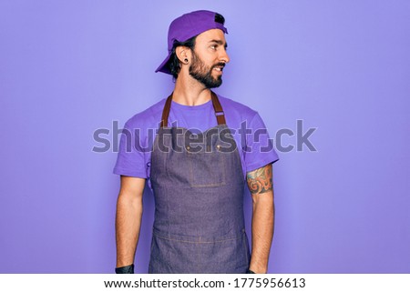 Young handsome hispanic man with bear wearing professional apron working as tattoo artist looking away to side with smile on face, natural expression. Laughing confident.