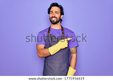 Young handsome hispanic clenaer man wearing housework apron and washing gloves cheerful with a smile of face pointing with hand and finger up to the side with happy and natural expression on face