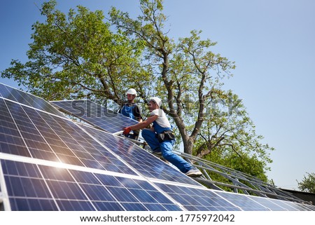 Two workers in protective helmet on steel platform installing heavy solar photo voltaic panel on green tree and blue sky background. Exterior solar panel system installation, dangerous job concept. Royalty-Free Stock Photo #1775972042