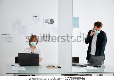 Pretty girl in medical mask and guy, talking on phone, work at computers in office at a safe distance from each other