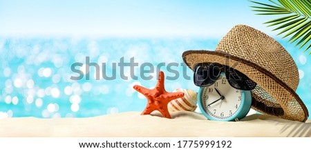 Summertime vacation concept. Time to relax. Last minute deals. Alarm clock with straw hat, starfish on the sand beach and sea background. Royalty-Free Stock Photo #1775999192