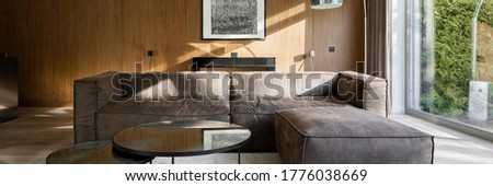 Panorama of stylish corner sofa with two modern coffee tables in living room with wooden and window walls