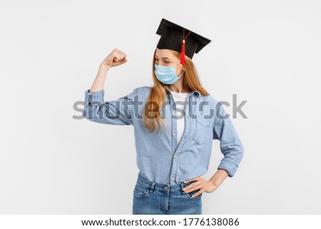 happy beautiful graduate girl, wearing a medical protective mask on her face and a graduation cap on her head, raises her hand and shows her muscles