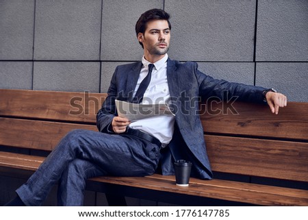 Daily news. Full length of a pleasant smiling businessman reading a newspaper with coffee while sitting on the bench.