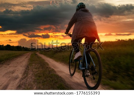 Girl on a bicycle at sunset. A cyclist in a helmet rides along a dirt path against the backdrop of a beautiful sky at sunset. Beautiful evening landscape.