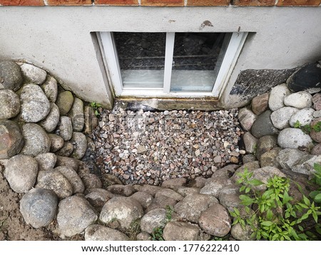 Photo showing recently fixed and reinstalled basement windows and window well surrounded by stones due to previous water accumulation inside of the pit. Royalty-Free Stock Photo #1776222410