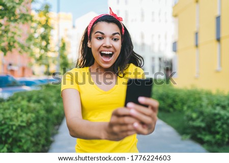 Surprised brunette woman in yellow t-shirt looking at smartphone screen shocked and excited with open mouth happy to win in online lottery.