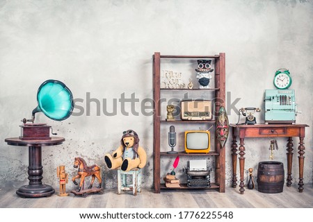 Antiques still life with old gramophone, Teddy bear, rocking horse, collection of media and writers devices, outdated cash register, clock front concrete wall background. Vintage style filtered photo