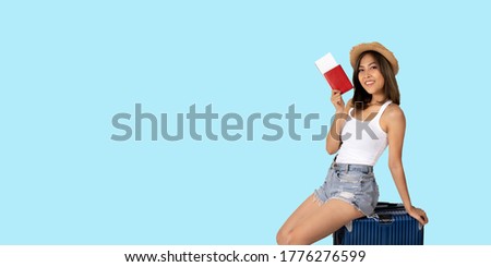 Panoramic of young woman going on vacation wearing hat and holding passport, sitting on luggage against plain blue background