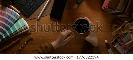 Overhead shot of female designer take a break with coffee on wooden table with painting tools and digital devices 