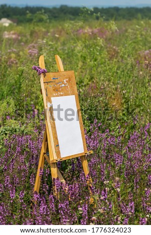 Wooden easel in a green field with purple flowers. White sheet of paper to create a picture.