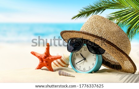 Summertime vacation concept. Time to relax. Alarm clock with straw hat, starfish on the sand beach and sea background. Royalty-Free Stock Photo #1776337625