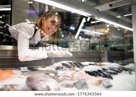 Supermarket worker arranging frozen fish for sale.Woman in working uniform selling fish in grocery store.  Royalty-Free Stock Photo #1776343364