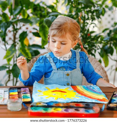 little toddler girl painting rainbow and sun with water colors during pandemic coronavirus quarantine disease. Children painting rainbows around the world . Happy child.