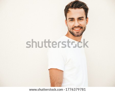 Portrait of handsome smiling stylish hipster lambersexual model.Man dressed in white T-shirt. Fashion male posing on the street background near wall in round sunglasses