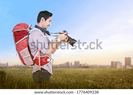 Asian man with a backpack holding the camera on the meadow field. World Photography Day