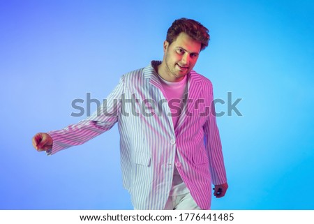 Young caucasian musician in casual dancing on gradient blue background in neon light. Concept of music, hobby, festival. Joyful party host, DJ, stand upper, dancer. Colorful portrait of artist.